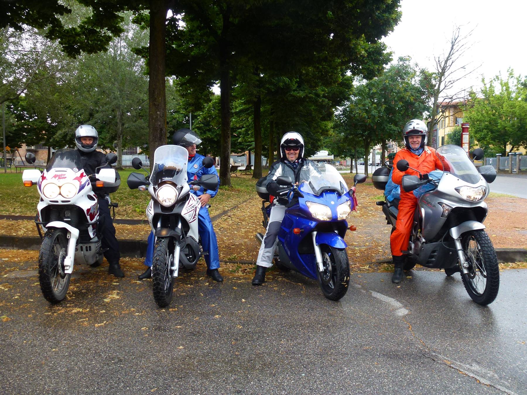 Quattro motociclisti con casco in sella a moto da viaggio, strada bagnata, alberi verdi