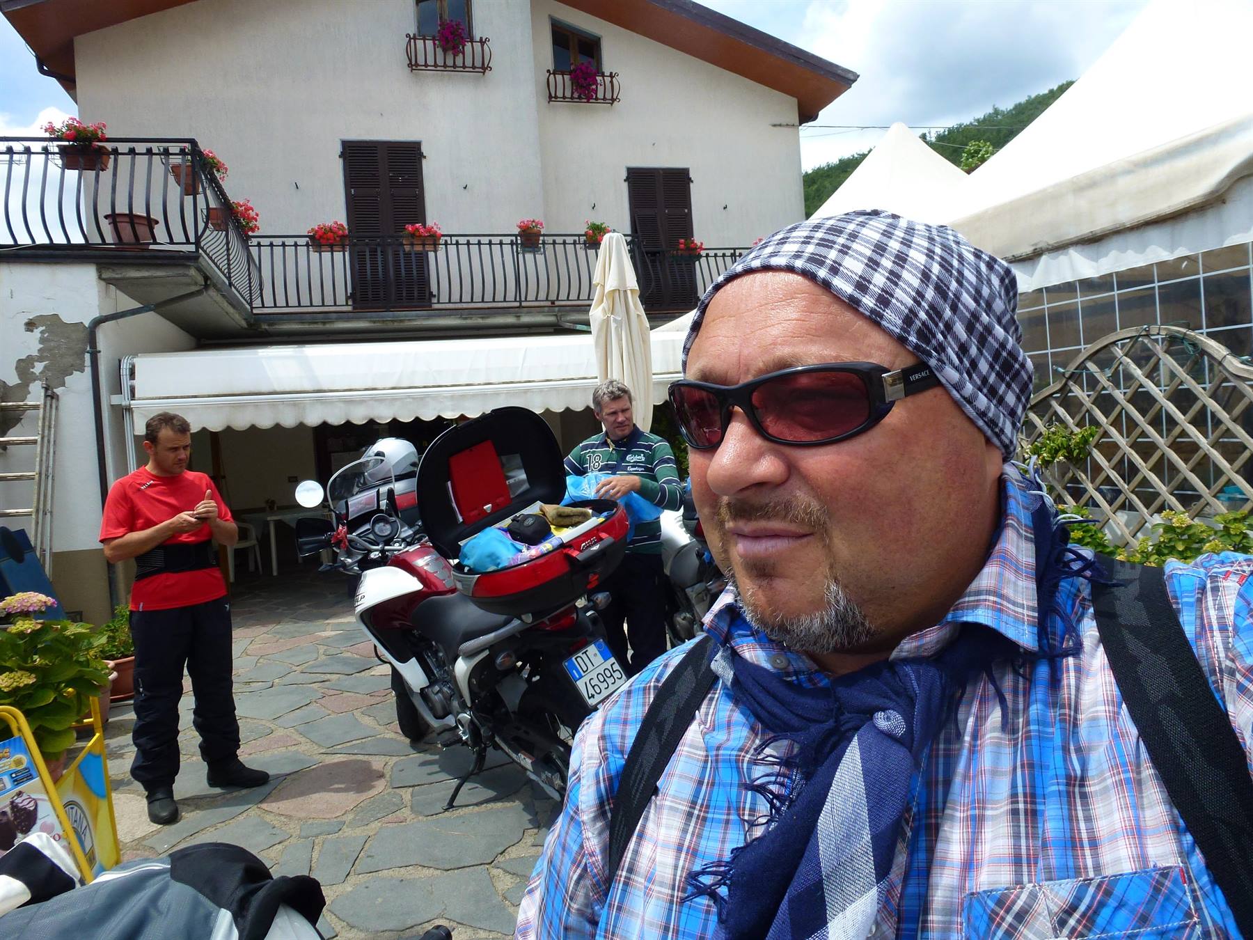 Motociclista con bandana e occhiali davanti a casa con balconi fioriti, moto caricate