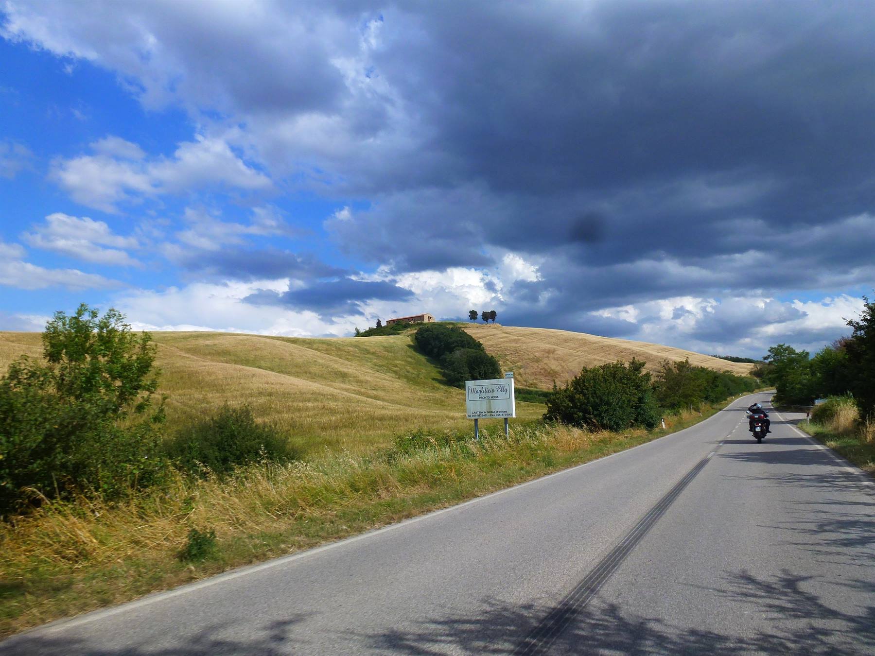 Motociclista su strada asfaltata tra colline coltivate e cielo nuvoloso