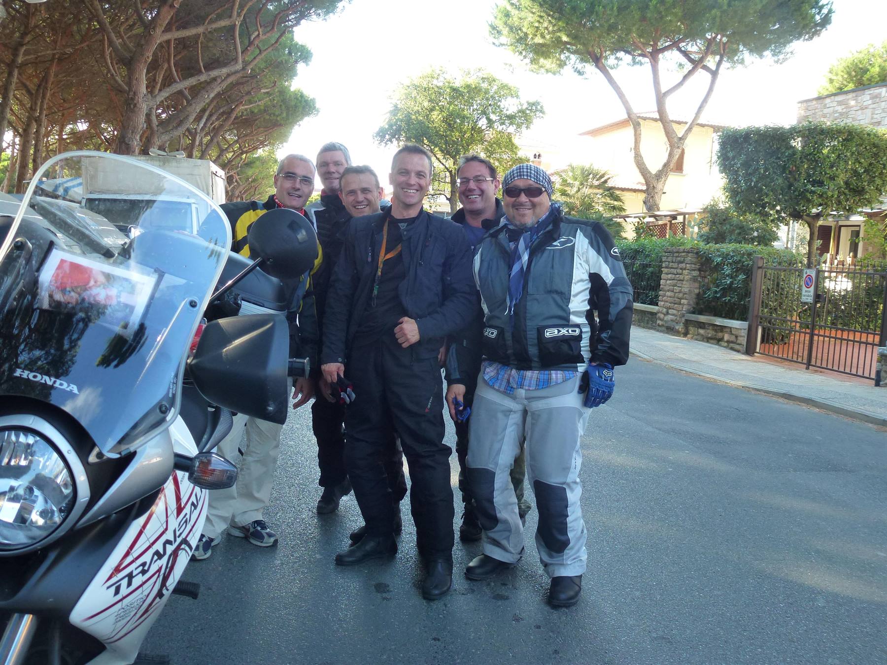 Cinque motociclisti in tuta accanto a una Honda Transalp, viale alberato al mattino
