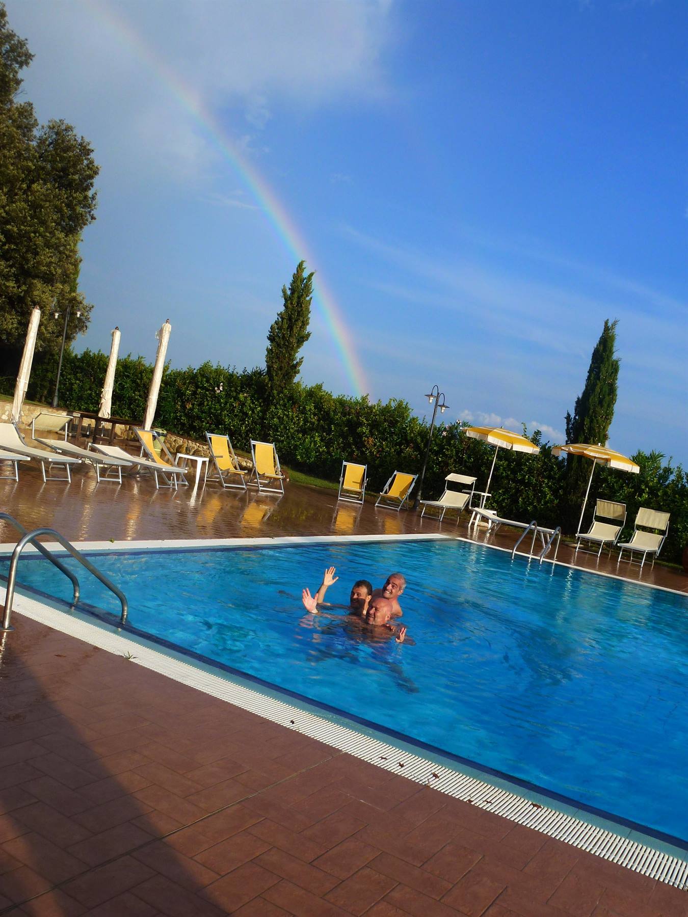 Due persone nuotano nella piscina con l'ombrelloni gialli e un arcobaleno