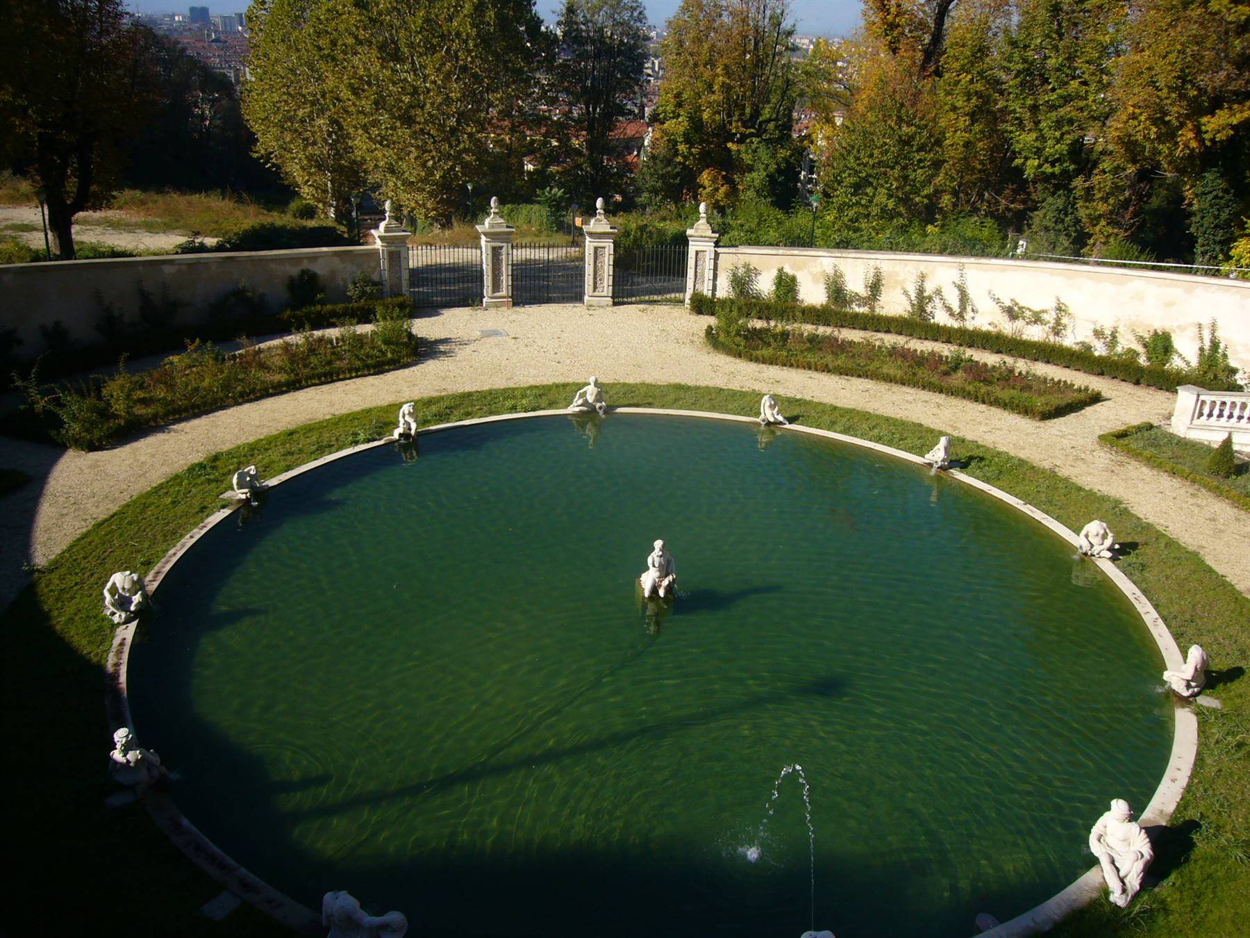 Vasca circolare verde con statue bianche e getto d'acqua, giardino barocco sotto il sole