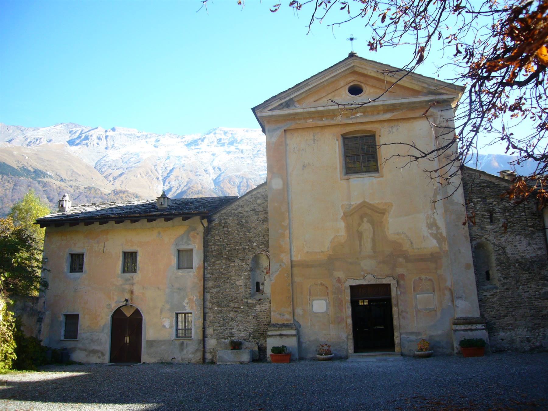 Chiesa antica con facciata ocra e case in pietra, montagne innevate sullo sfondo