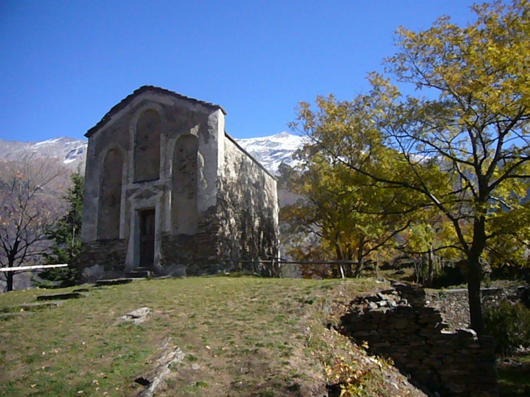 Antica chiesa in pietra tra alberi autunnali, monti innevati sullo sfondo, cielo azzurro