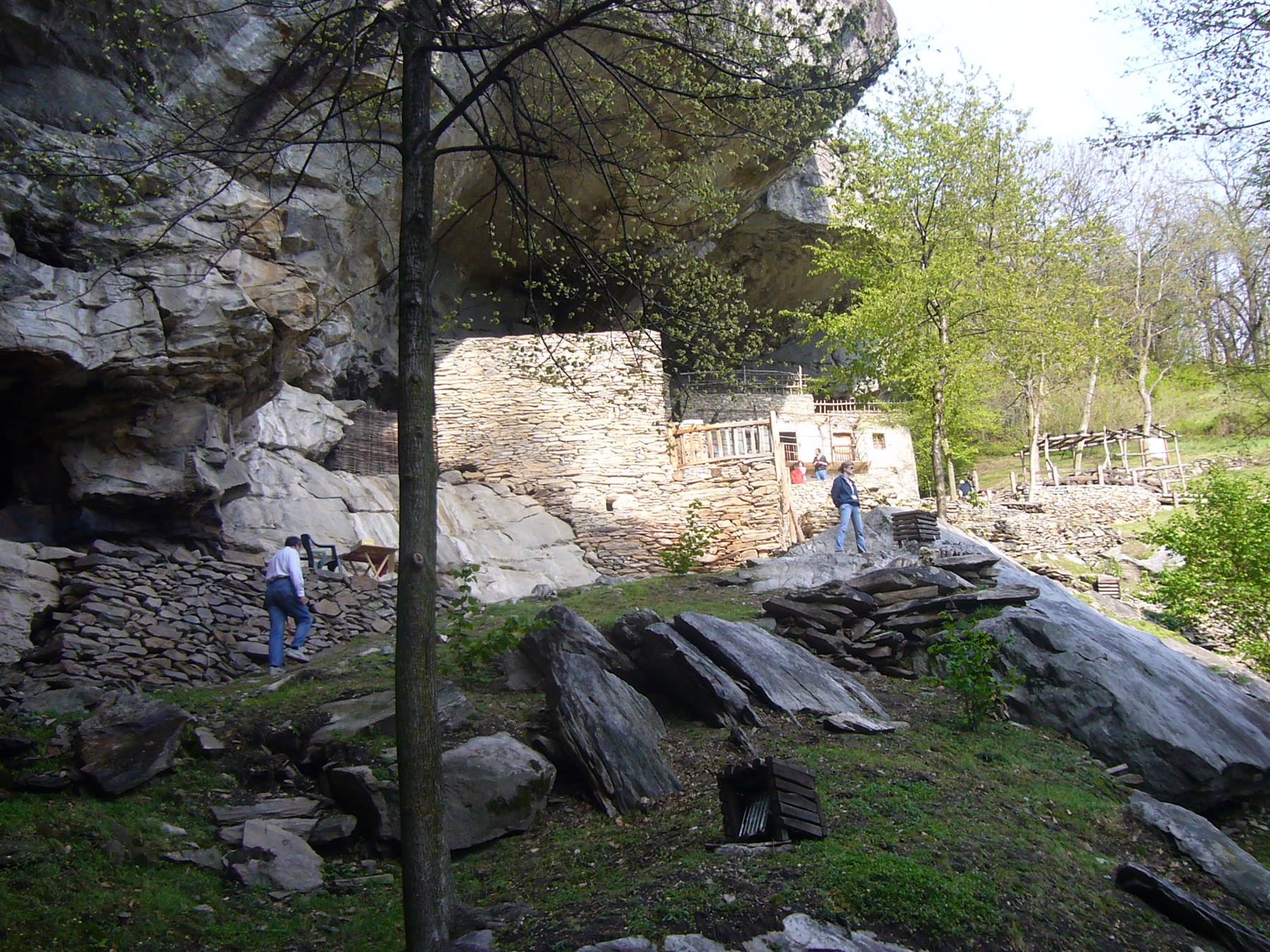 Turisti vicino a muretti in pietra sotto un'enorme roccia boscosa