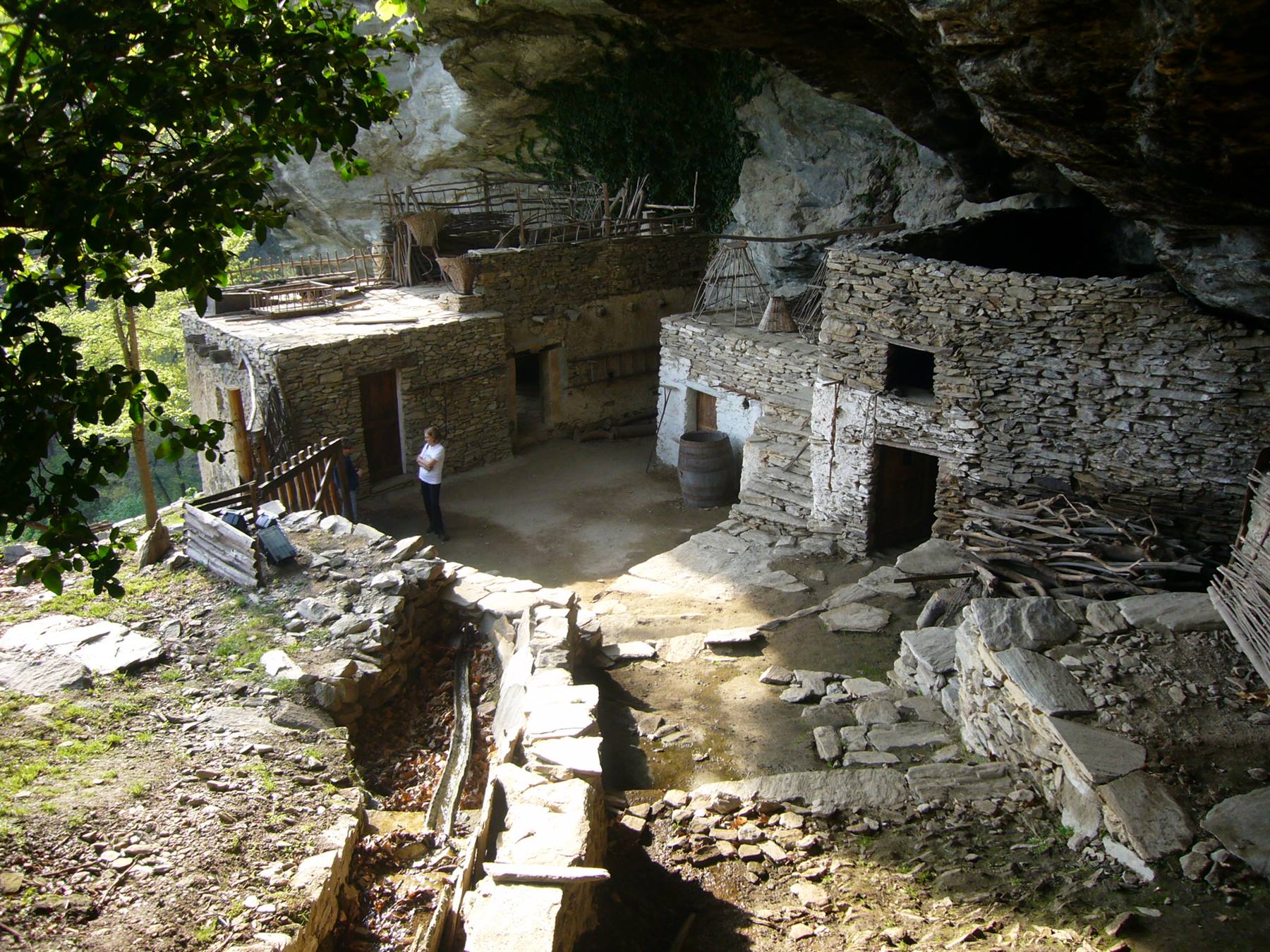 Antichi ricoveri in pietra a secco sotto una sporgenza rocciosa, una visitatrice osserva