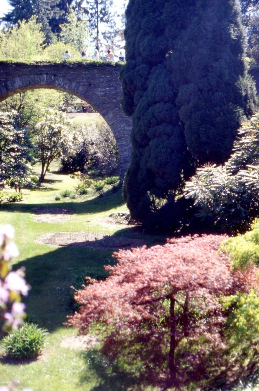 Visitatori attraversano un ponte ad arco in pietra sopra giardini al sole