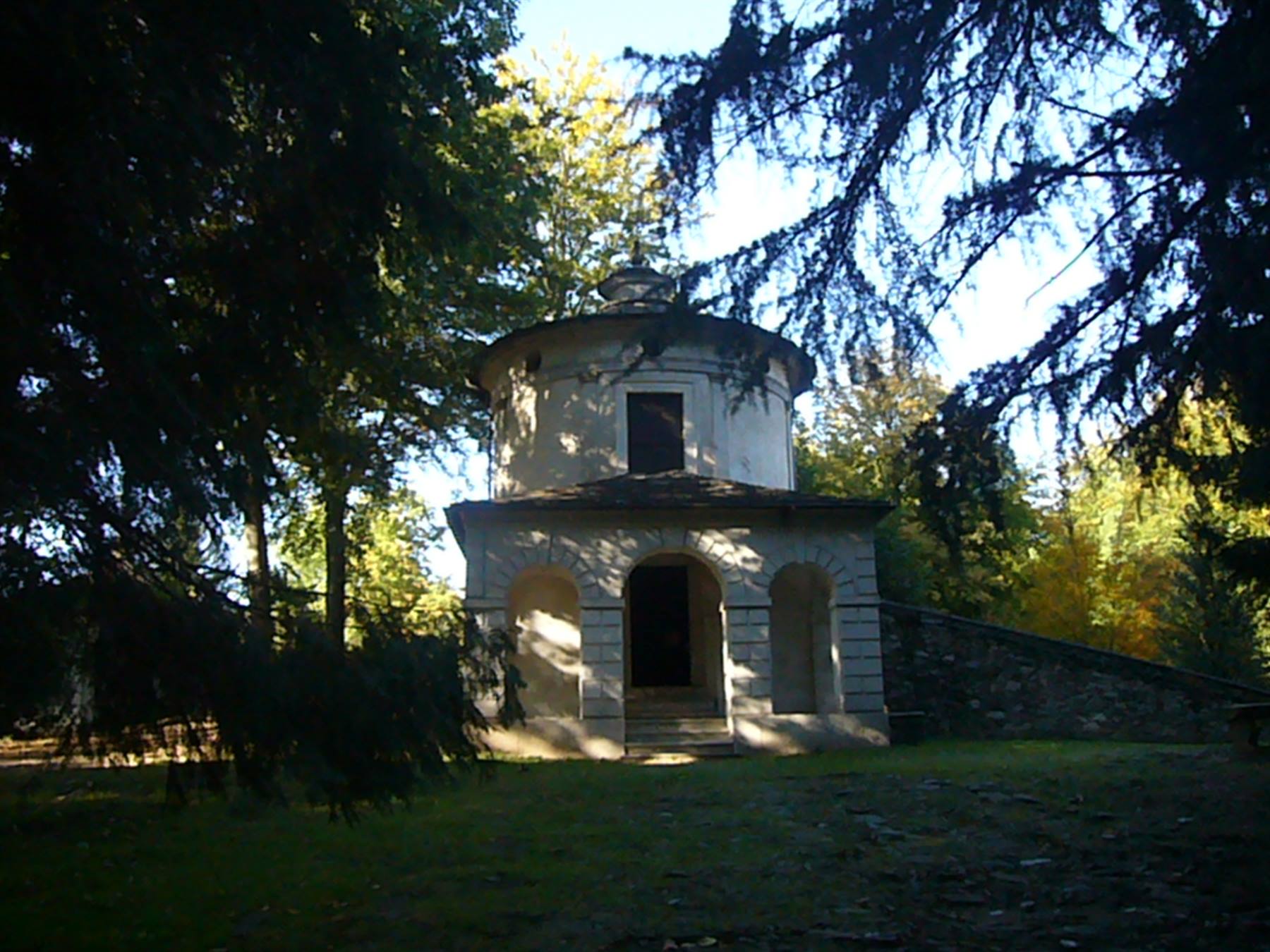 Cappella circolare in pietra chiara con archi, tra alberi al sole
