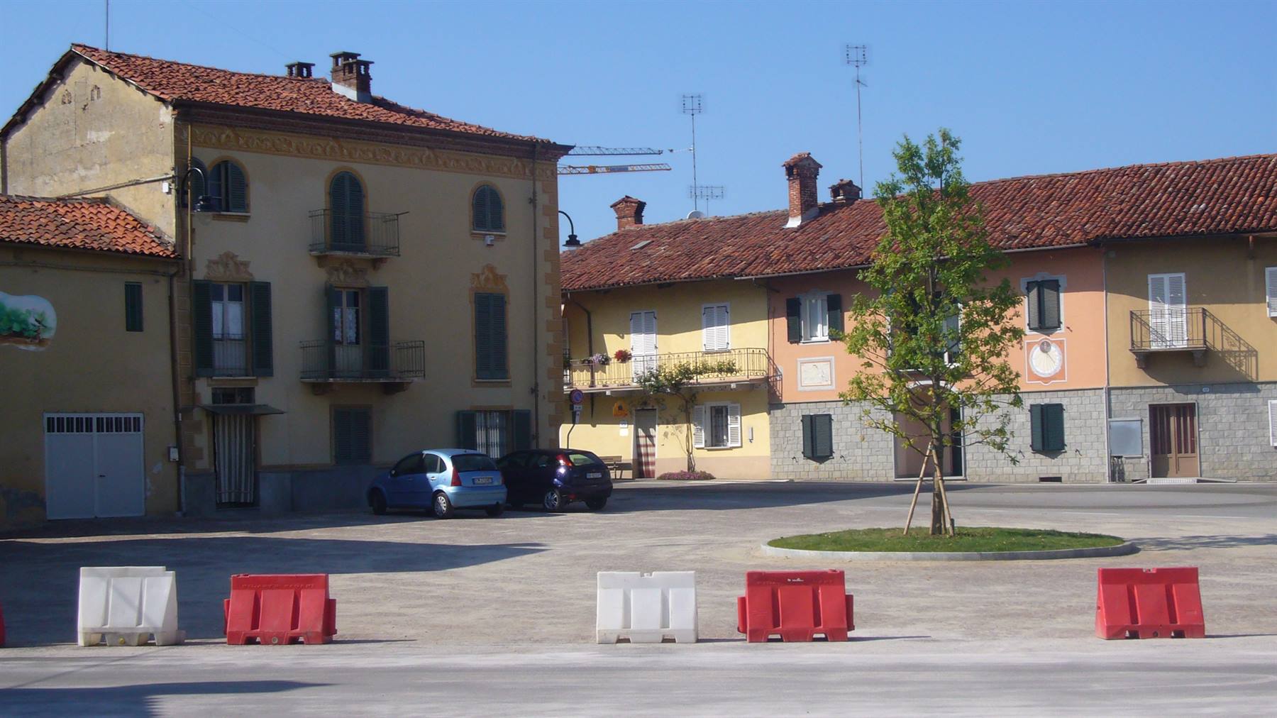 Piazza di paese con case colorate, tetti in cotto, auto parcheggiate e barriere rosse