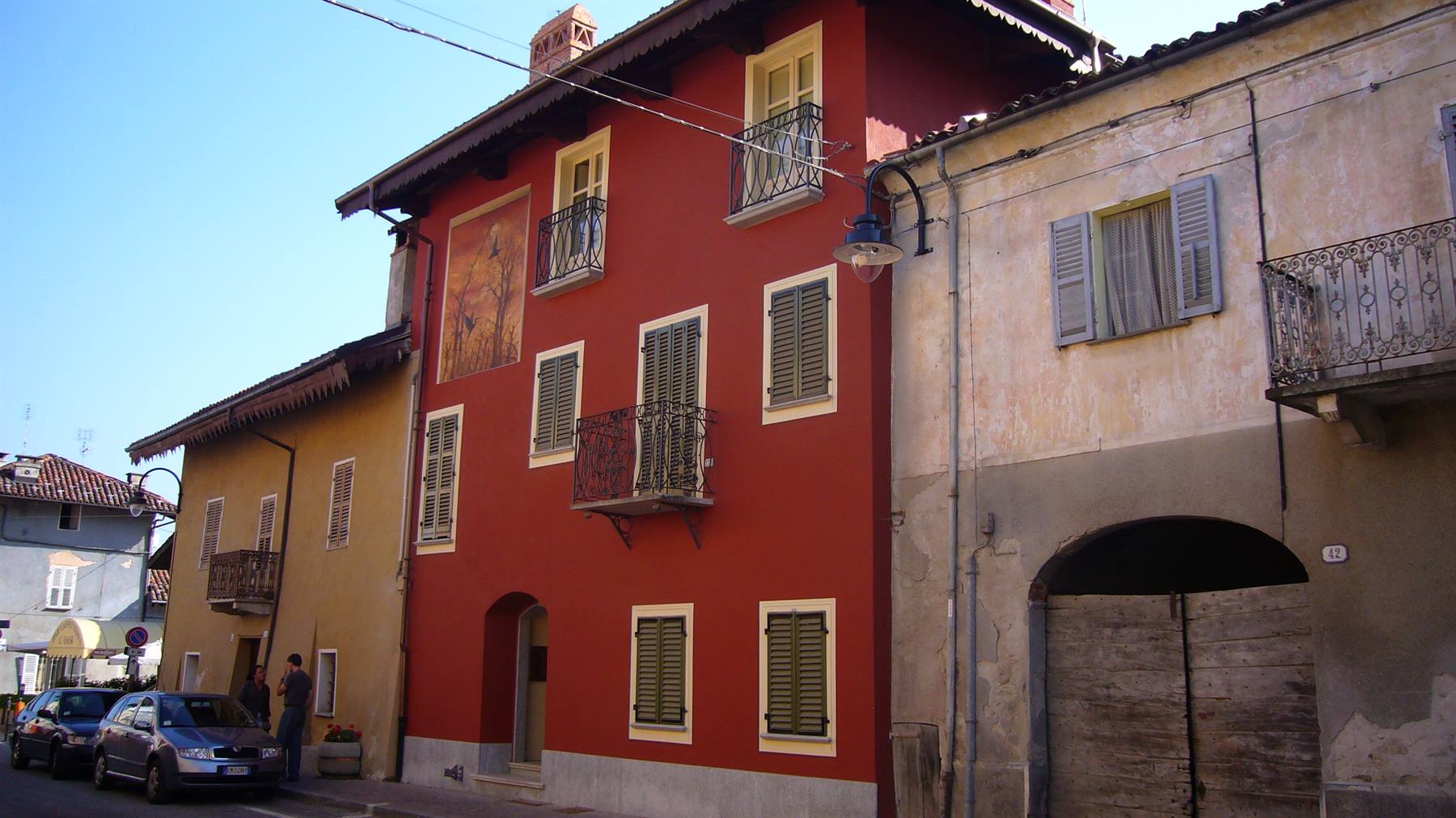 Casa rossa con affresco, balconi in ferro e persiane verdi lungo la strada