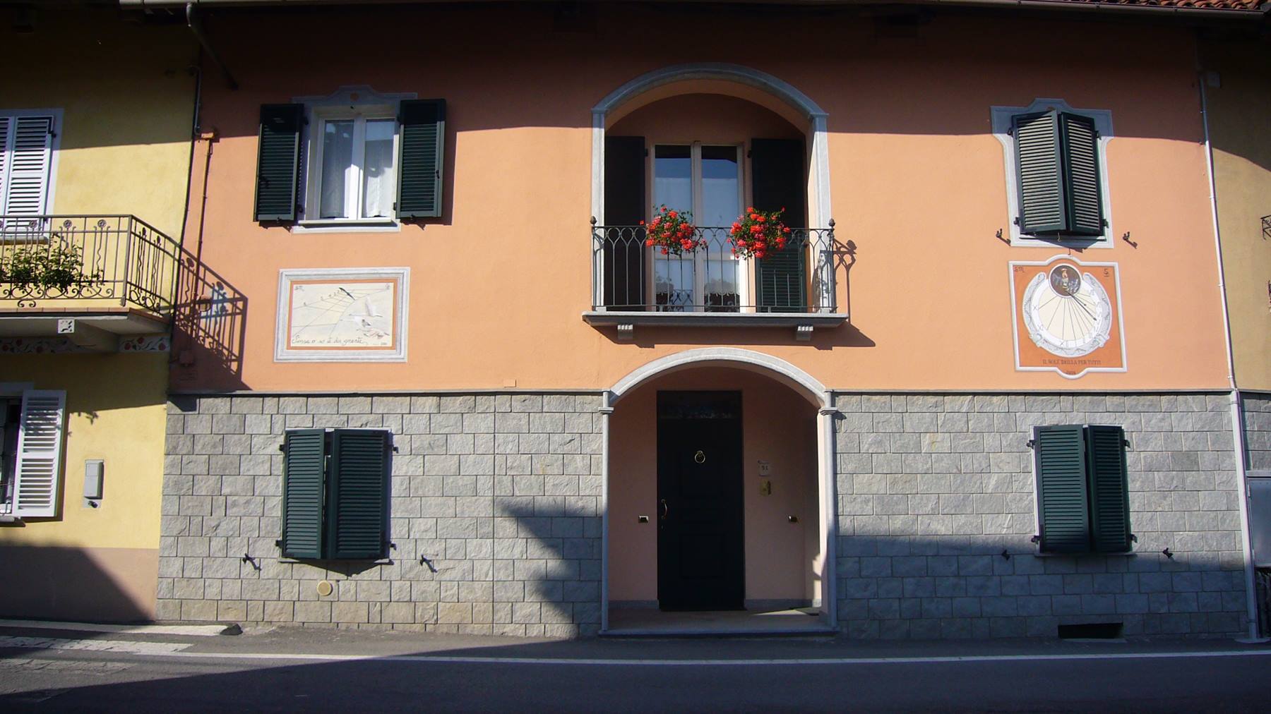 Facciata rosa con meridiana affrescata, persiane verdi e gerani sul balcone