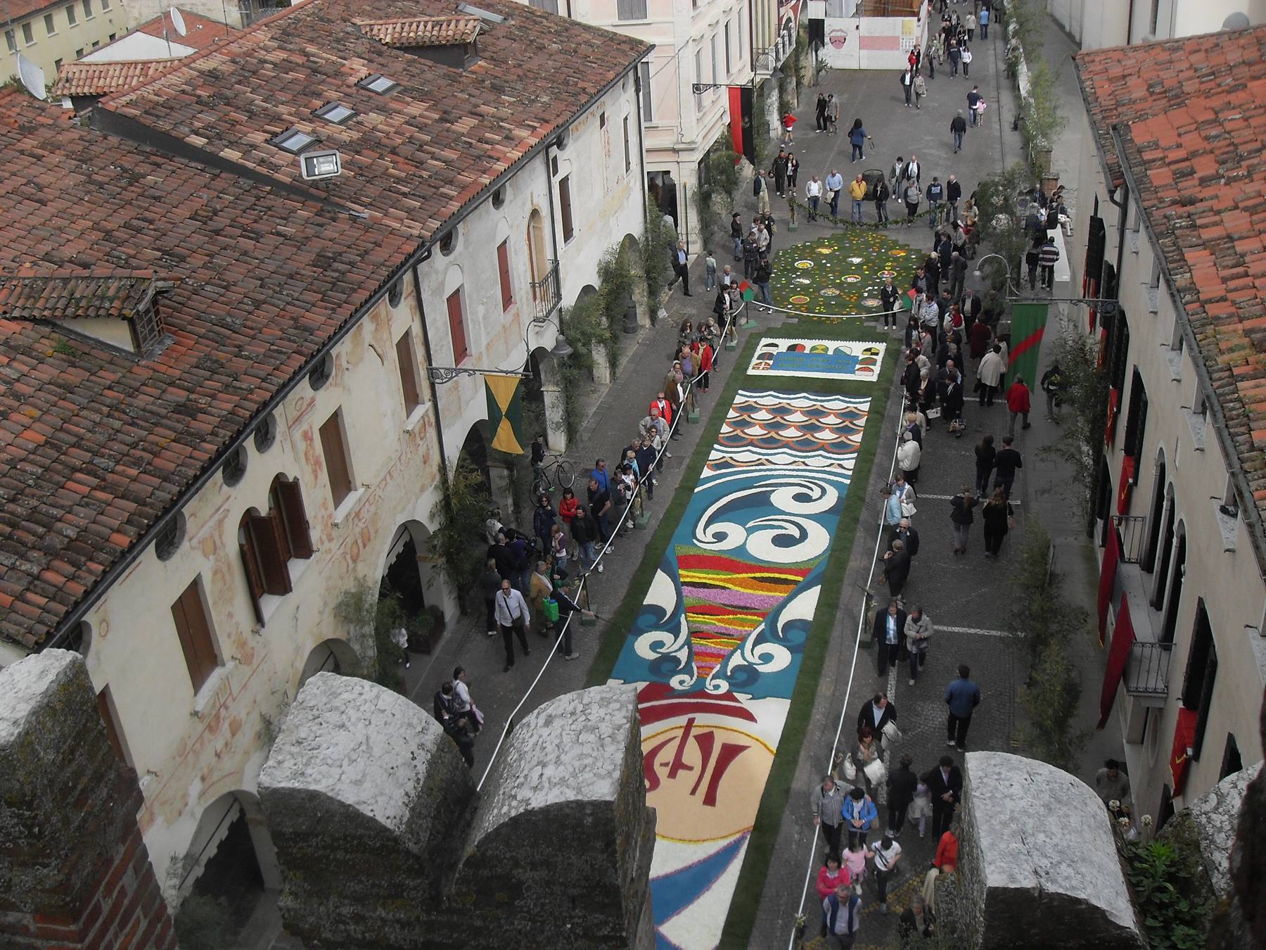 Infiorata: tappeto di fiori colorato in strada, folla tra case con tetti in coppi