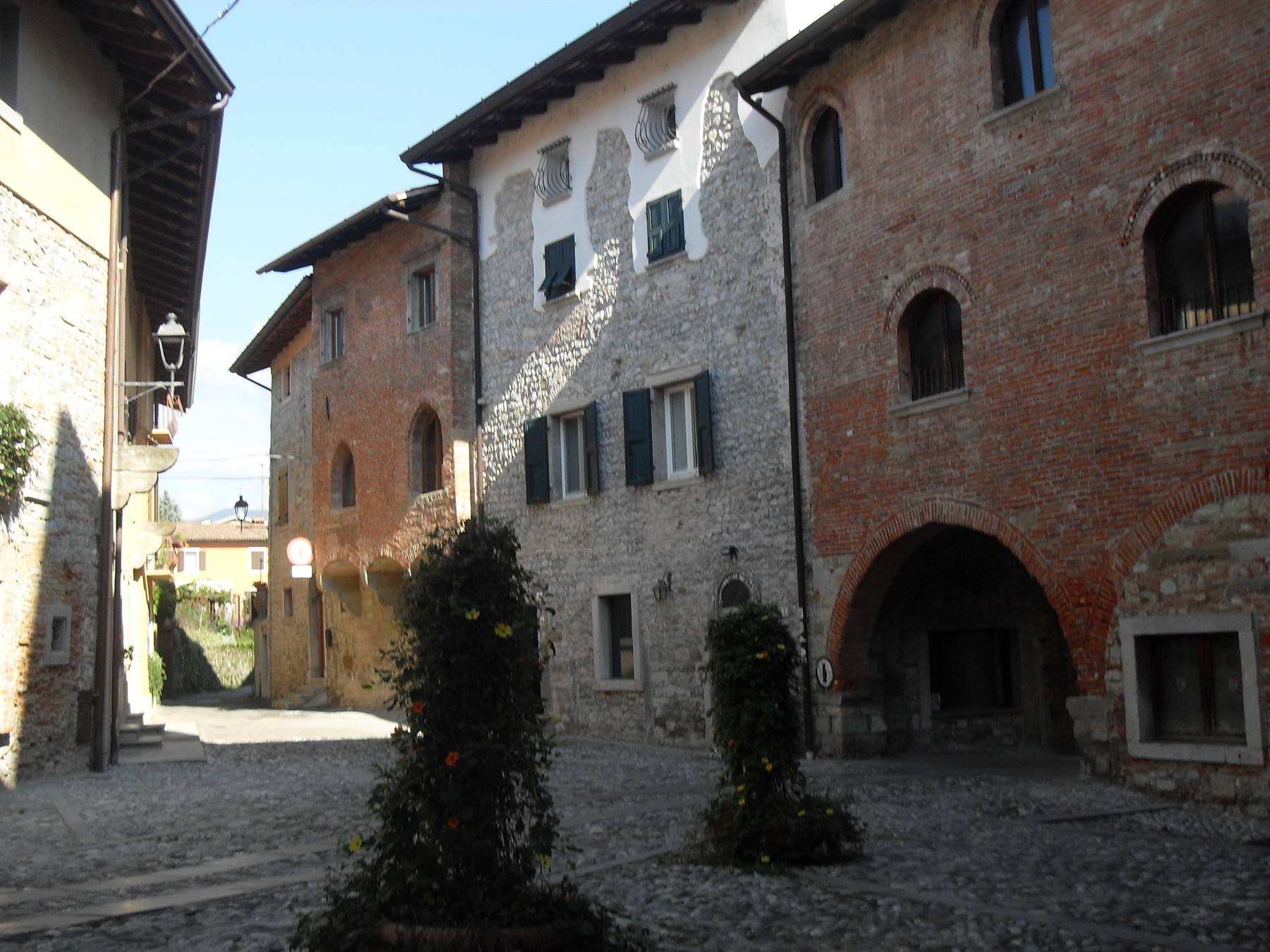 Piazzetta in ciottoli con case in pietra e mattoni, arcate e cespugli fioriti