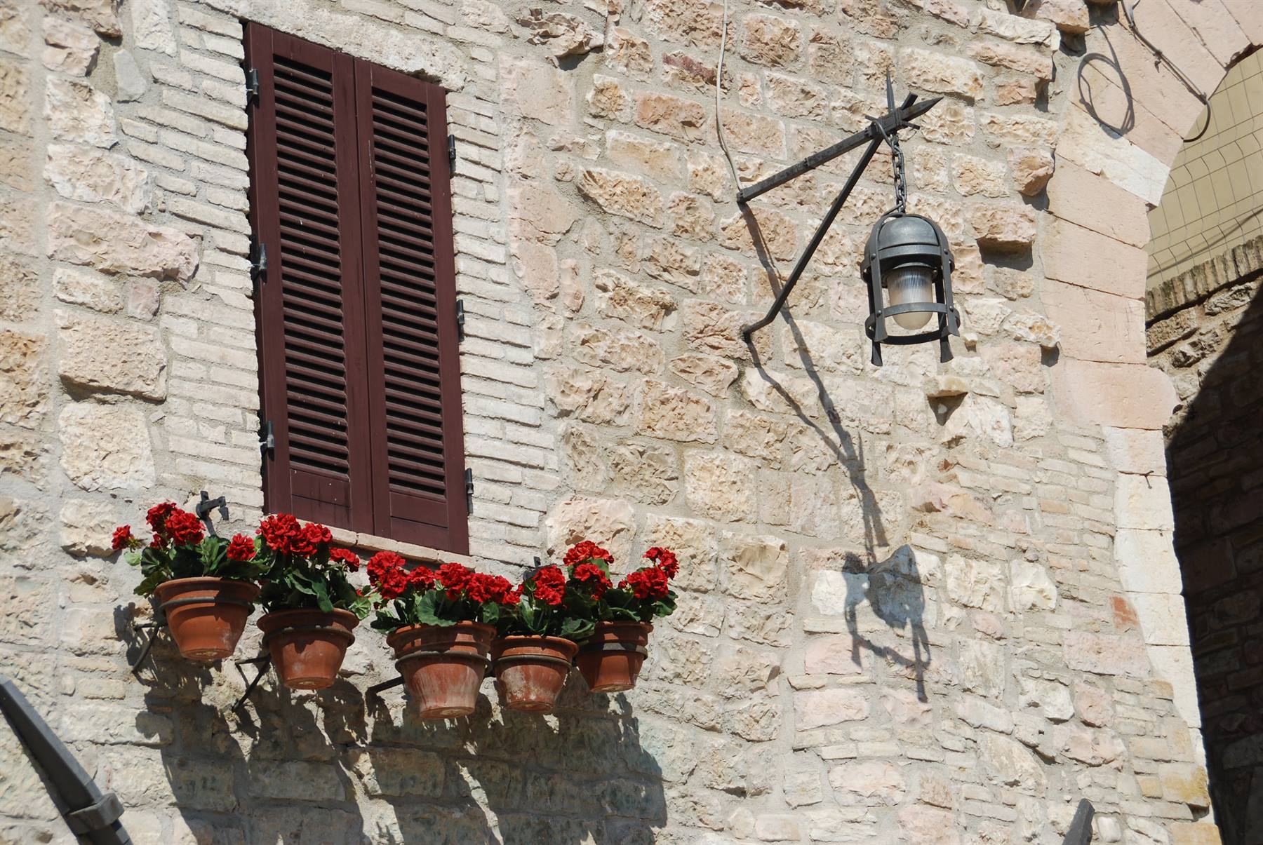 Muro di pietra con persiana marrone, vasi di gerani rossi e lanterna di ferro