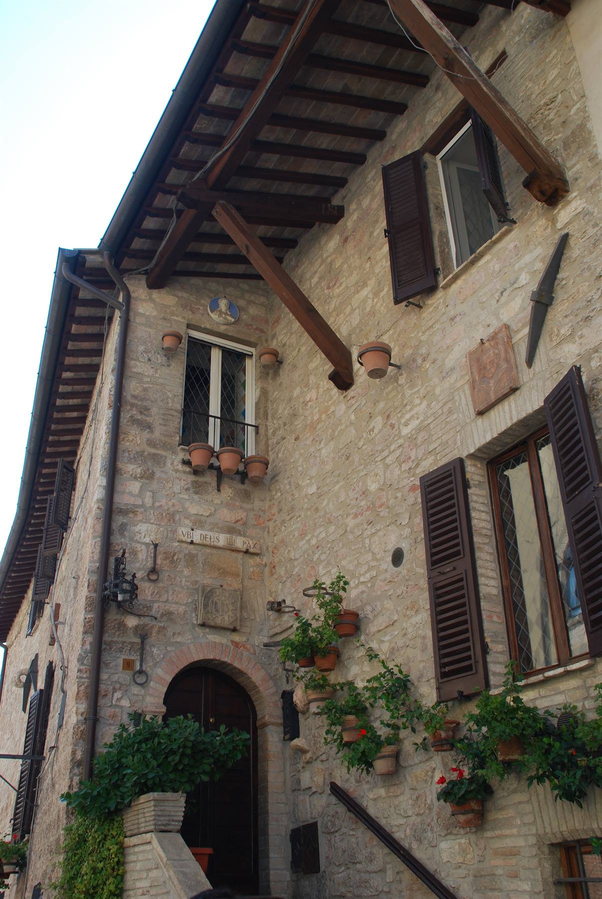 Casa in pietra con travi di legno, persiane scure e vasi di fiori appesi, Assisi