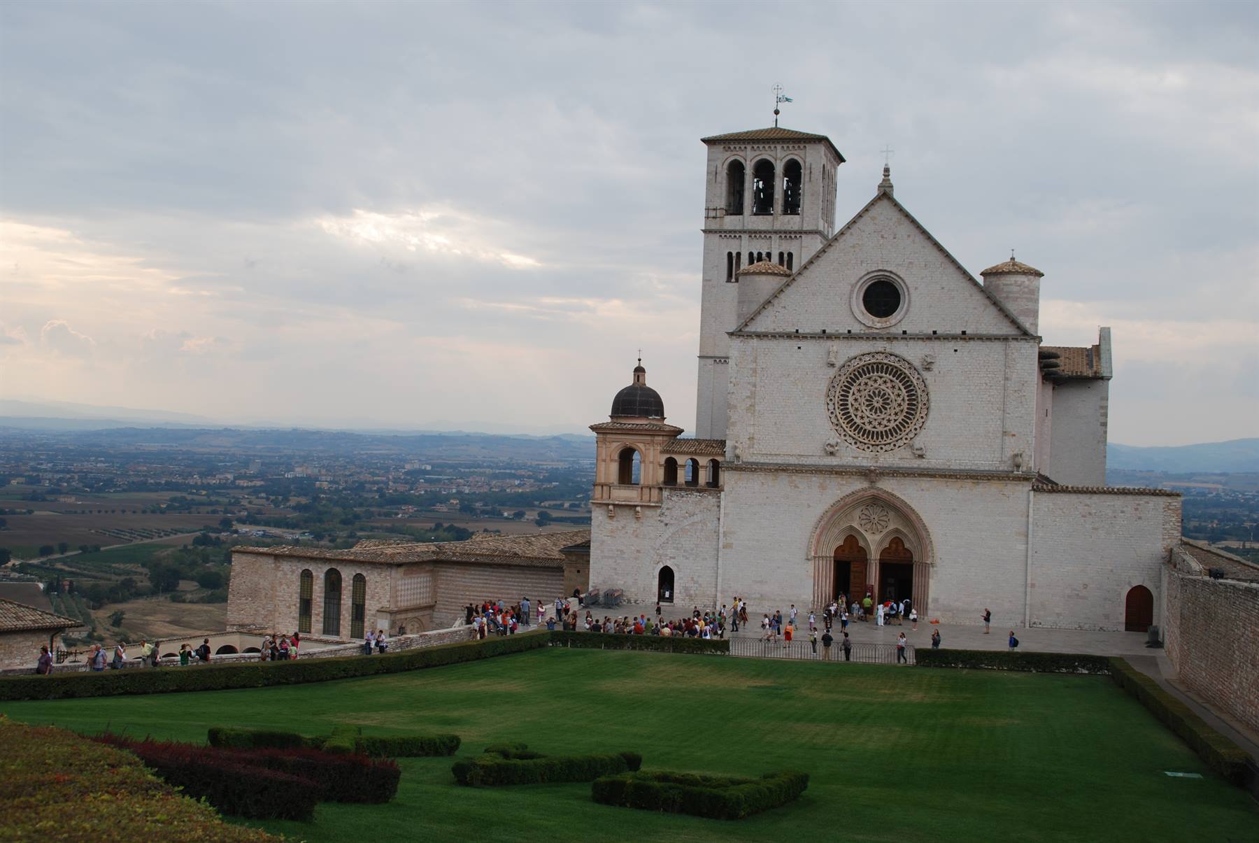 Basilica di San Francesco ad Assisi, facciata bianca con rosone, visitatori sul prato