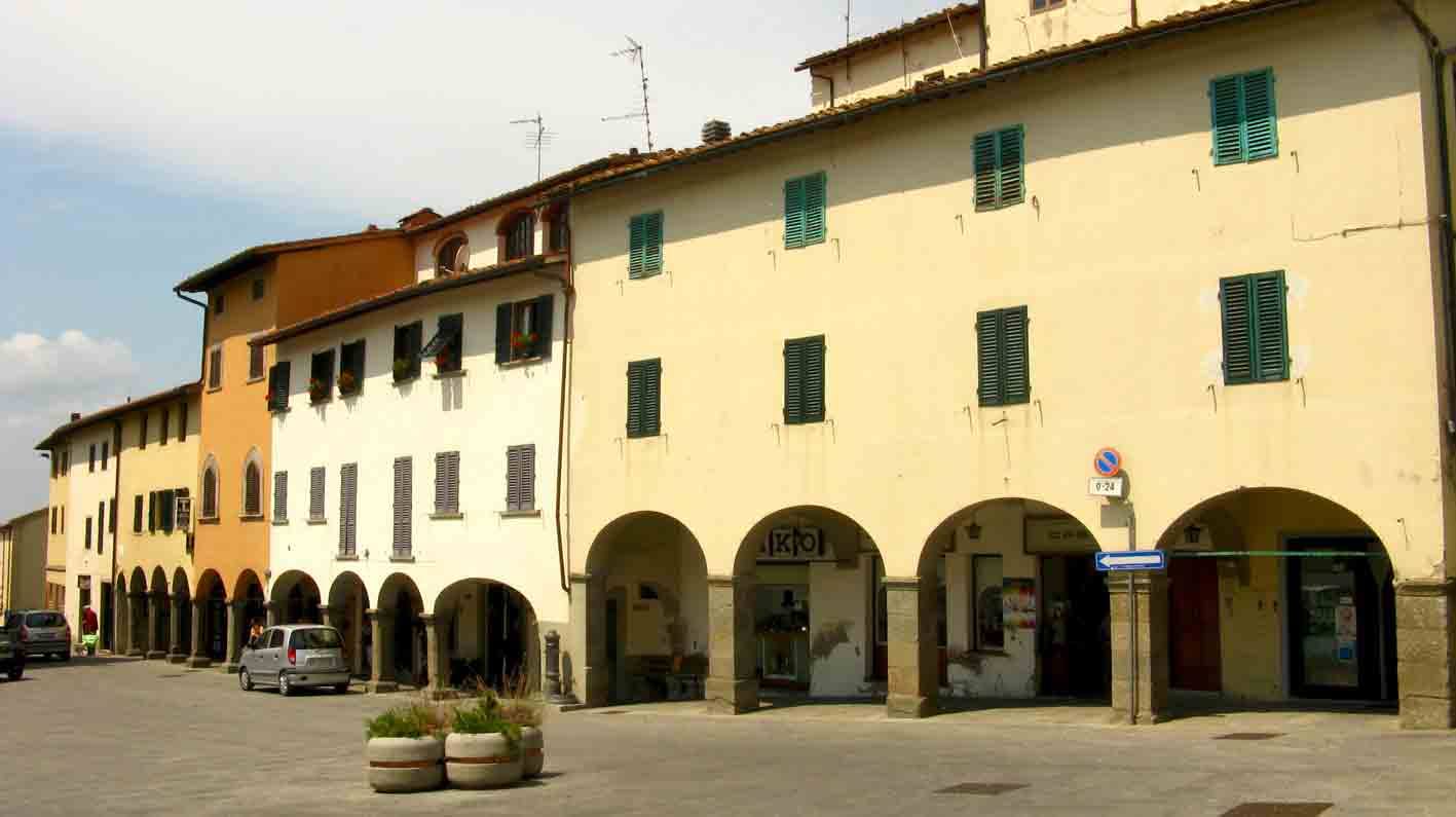 Piazza con edifici porticati ocra e gialli, persiane verdi, automobili e fioriere in pietra