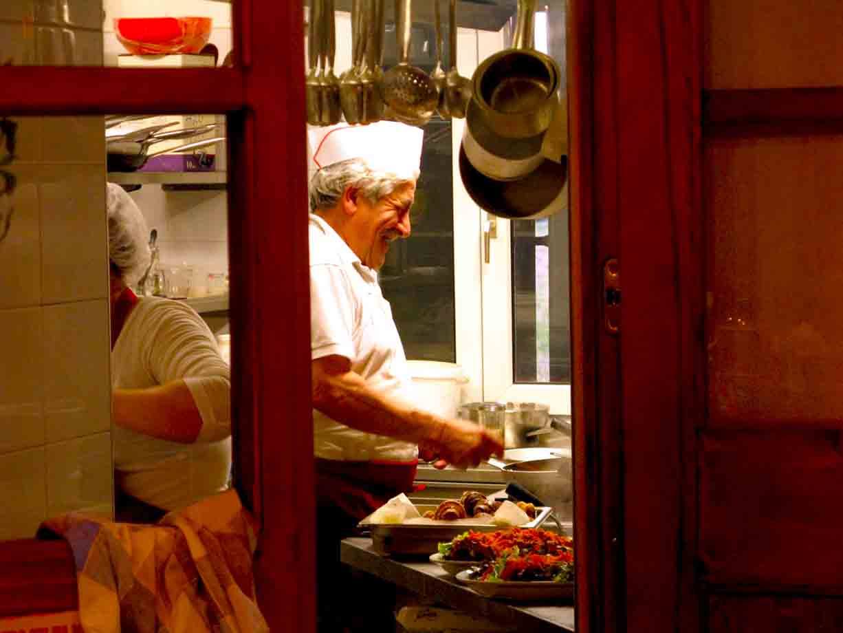 Cuoco con cappello bianco impiatta nel ristorante, visto da una porta rossa