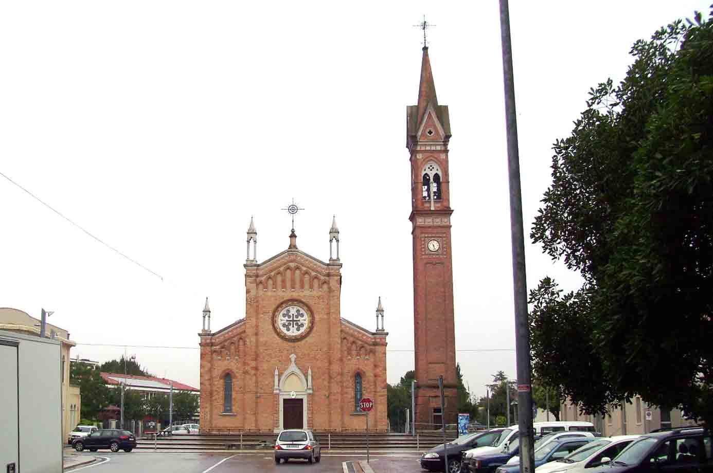 Chiesa in mattoni con rosone e campanile, piazza con auto ferme sotto cielo grigio