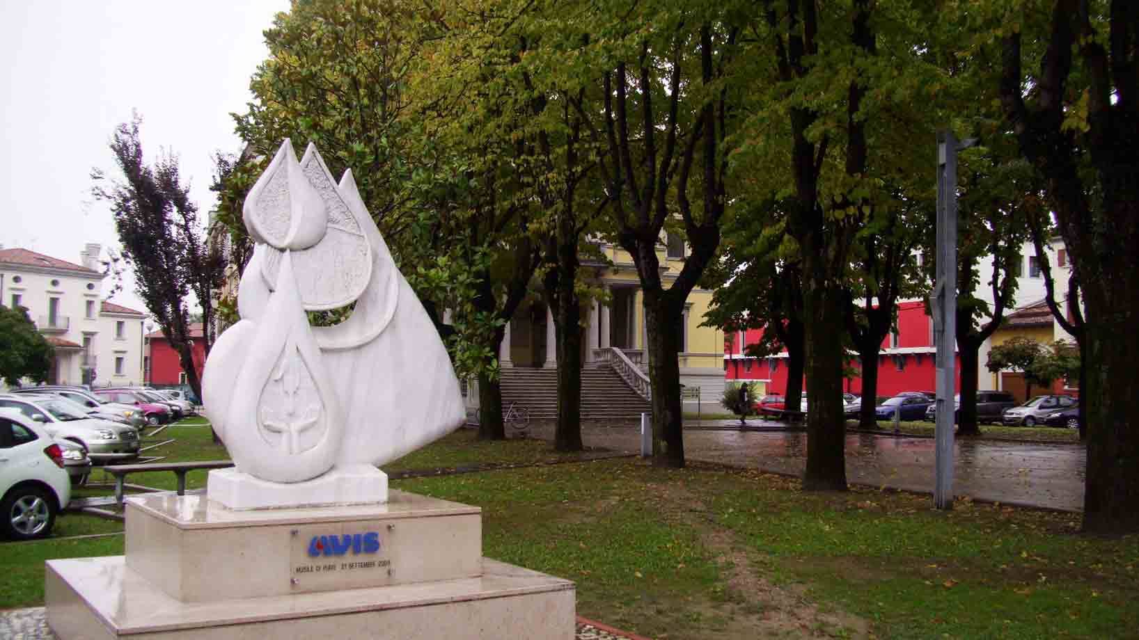 Scultura in marmo bianco a forma di goccia su piazza alberata, davanti a edificio rosso