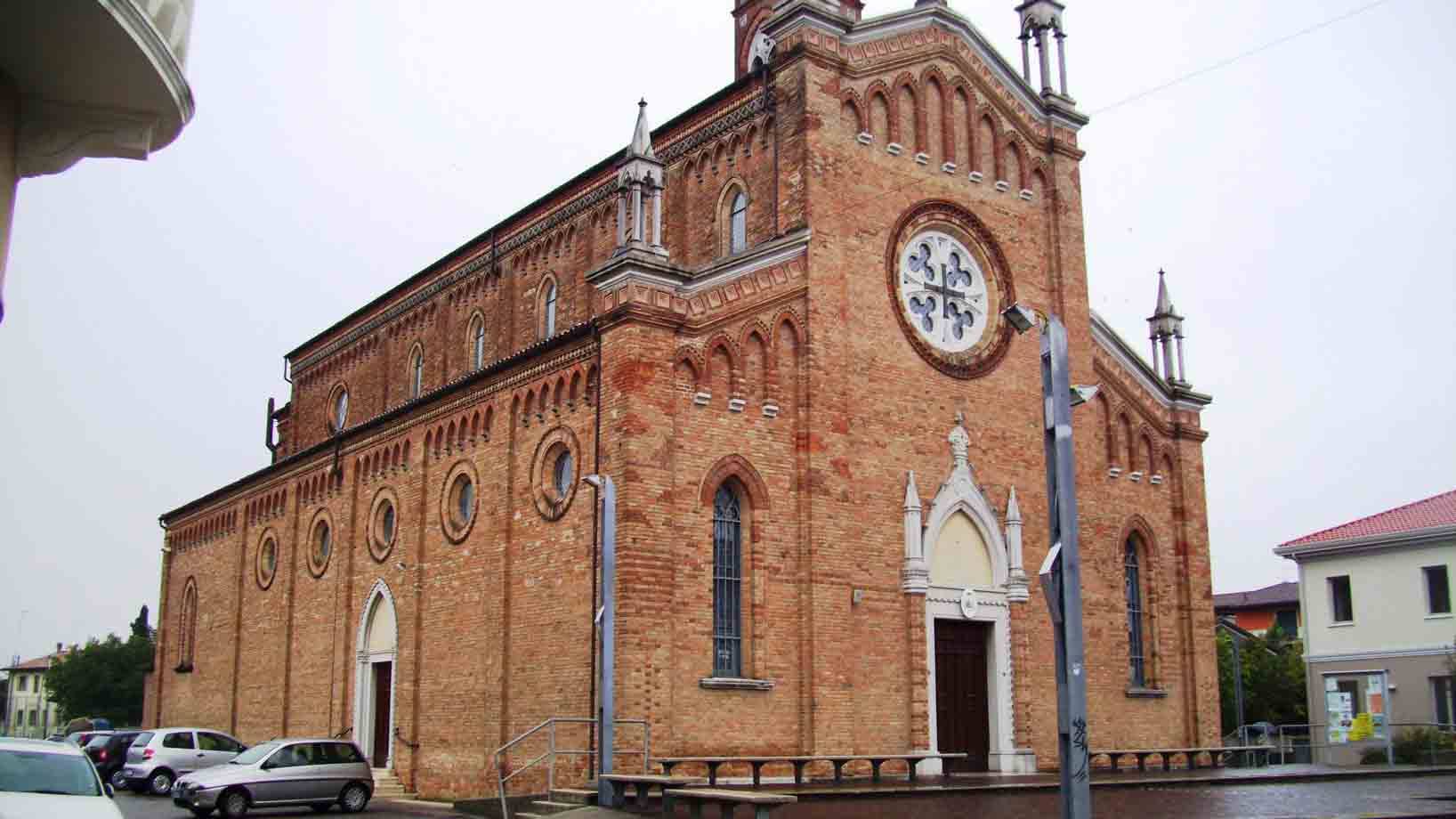 Chiesa in mattoni con rosone e guglie sotto cielo grigio, auto parcheggiate davanti