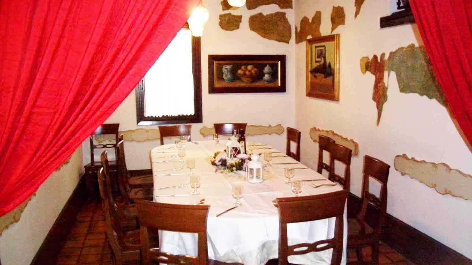 Sala di ristorante con tavola imbandita, tovaglia bianca, sedie in legno e tende rosse