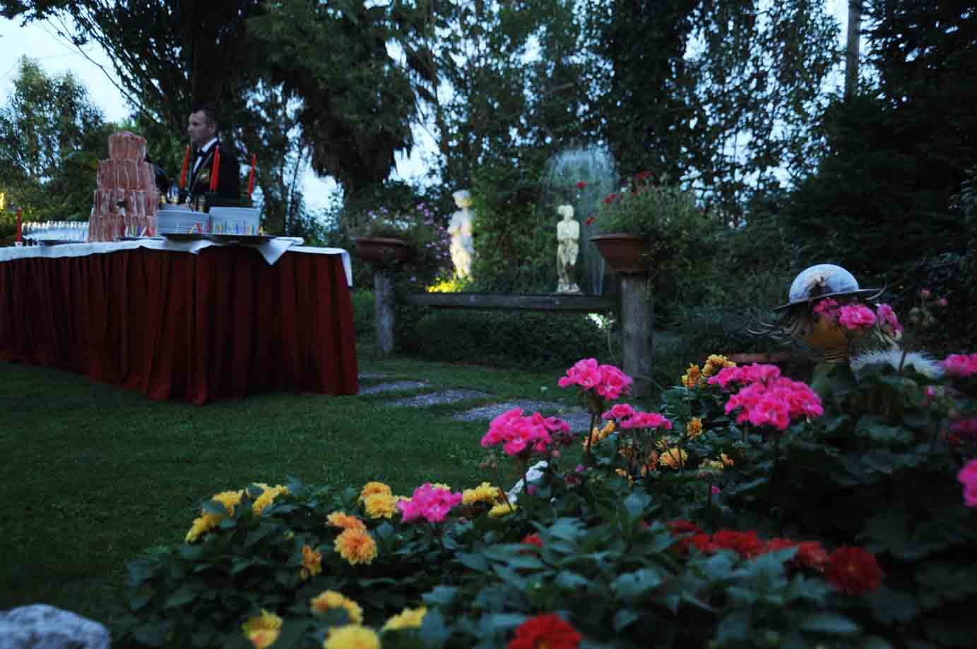 Torta nuziale a più piani sul tavolo imbandito, fiori rosa e statue nel giardino crepuscolare