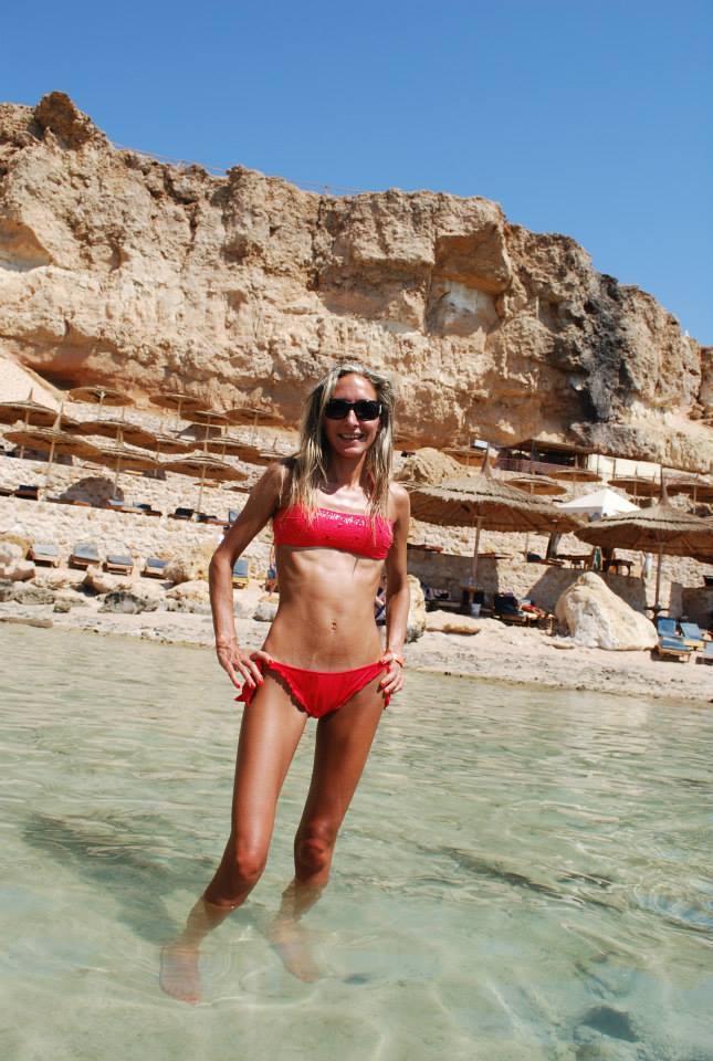 Donna in bikini rosso e occhiali da sole in acqua trasparente davanti a scogliera sabbiosa con ombrelloni