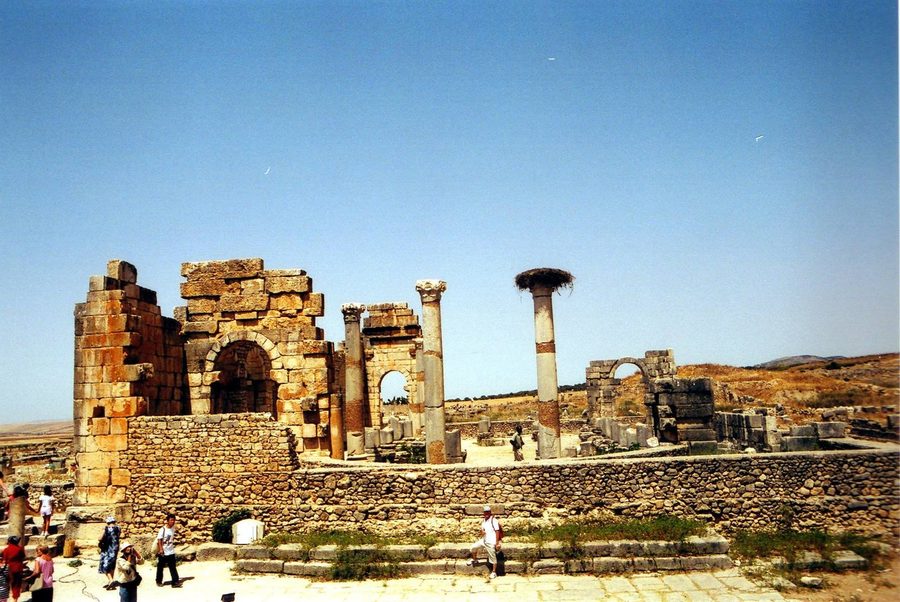 Rovine romane di Volubilis: colonne e archi di pietra sotto cielo azzurro, turisti camminano