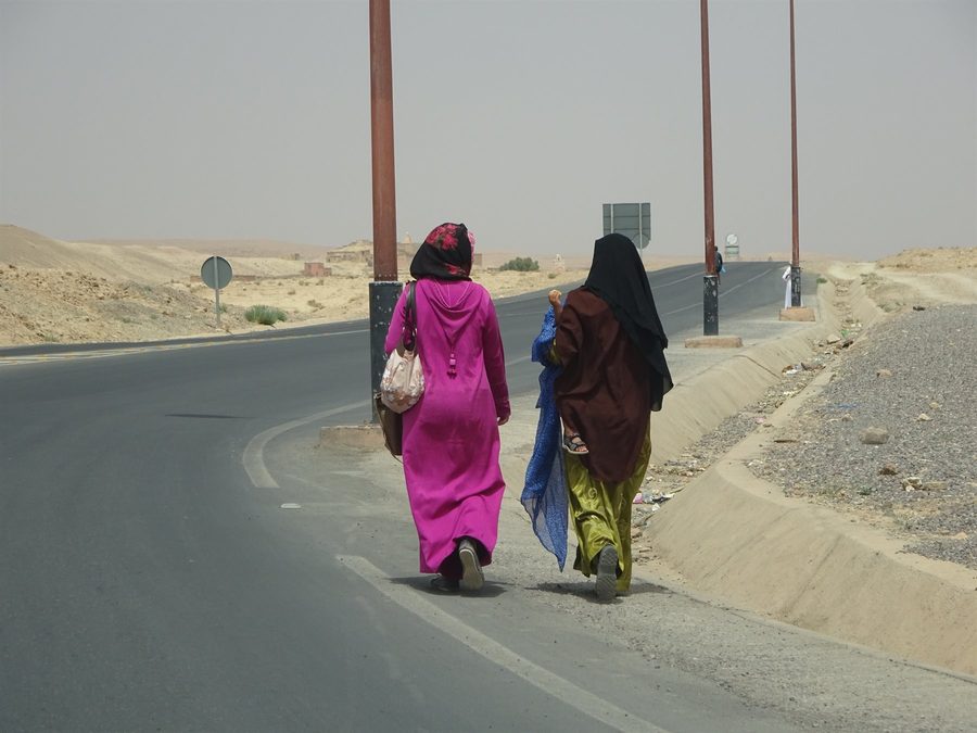 Donne in djellaba camminano lungo l'asfalto tra sabbia e lampioni a Merzouga