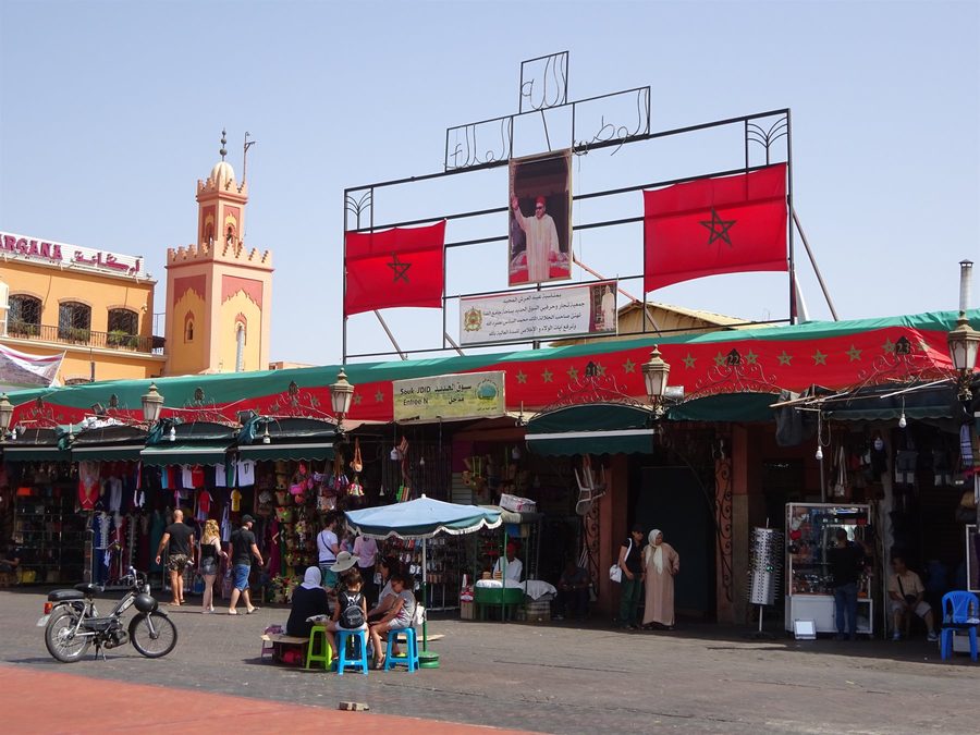 Piazza Jemaa el-Fna a Marrakech: bandiere marocchine, ritratto del re, minareto e bancarelle