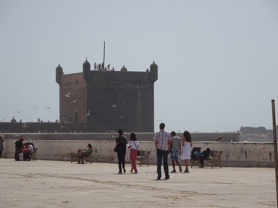 Turisti camminano verso la Sqala du Port a Essaouira, gabbiani nel cielo nebbioso